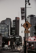 A bustling city street at dusk with several tall buildings, including one displaying a sign with the letters 'C&C'. The street is busy with cars and various illuminated signs and billboards. A pedestrian crossing sign is prominently displayed in the foreground, and banners are hung from streetlights.