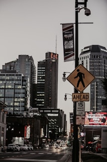 A bustling city street at dusk with several tall buildings, including one displaying a sign with the letters 'C&C'. The street is busy with cars and various illuminated signs and billboards. A pedestrian crossing sign is prominently displayed in the foreground, and banners are hung from streetlights.