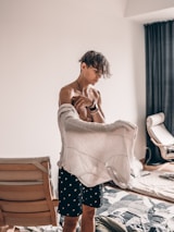 A person stands in a casually furnished room, holding a sweater in their hand. They appear to be wearing patterned shorts and are surrounded by a couple of wooden chairs and a mattress on the floor. The space is minimally decorated with a curtain in the background.