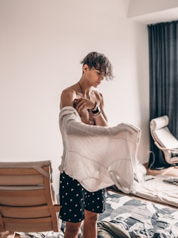 A person stands in a casually furnished room, holding a sweater in their hand. They appear to be wearing patterned shorts and are surrounded by a couple of wooden chairs and a mattress on the floor. The space is minimally decorated with a curtain in the background.