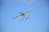 A small aerobatic airplane flies in the sky with a clear blue backdrop. The aircraft is primarily white with green and black accents.