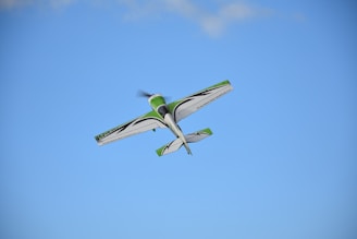 A small aerobatic airplane flies in the sky with a clear blue backdrop. The aircraft is primarily white with green and black accents.