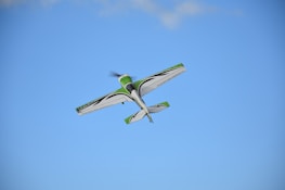 A small aerobatic airplane flies in the sky with a clear blue backdrop. The aircraft is primarily white with green and black accents.