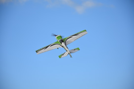 A small aerobatic airplane flies in the sky with a clear blue backdrop. The aircraft is primarily white with green and black accents.