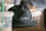 A black cap with a label that reads 'Build - Play', sitting on a wooden surface. In the background, there are blurred elements, including a clear plastic cup with a green straw and other indistinct objects.