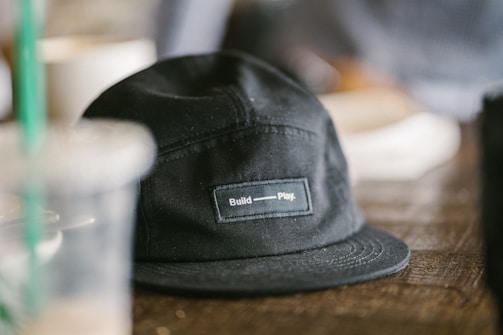 A black cap with a label that reads 'Build - Play', sitting on a wooden surface. In the background, there are blurred elements, including a clear plastic cup with a green straw and other indistinct objects.
