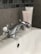 A chrome faucet mounted on a white porcelain sink, with a hint of a tiled wall in the background. Two tubes of skincare products are partially visible, positioned behind the faucet.