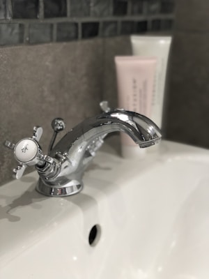 A chrome faucet mounted on a white porcelain sink, with a hint of a tiled wall in the background. Two tubes of skincare products are partially visible, positioned behind the faucet.