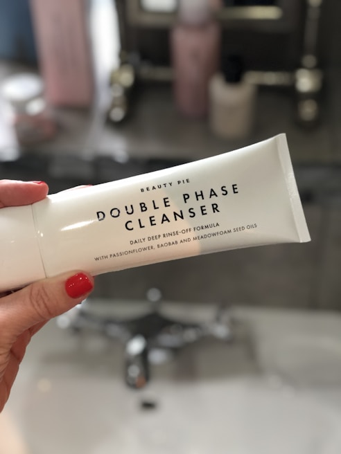A person with red nail polish is holding a white tube labeled 'Double Phase Cleanser' by Beauty Pie. The background is a bathroom with a sink and blurred toiletries.