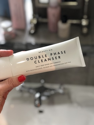 A person with red nail polish is holding a white tube labeled 'Double Phase Cleanser' by Beauty Pie. The background is a bathroom with a sink and blurred toiletries.