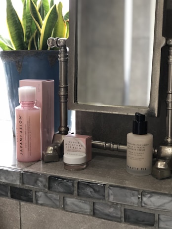 On a bathroom counter, there is a collection of cosmetic products including a pink bottle with the label 'JapanFusion', a small jar labeled 'Super Moisture Lip Balm', and a beige bottle labeled 'Beauty Pie'. A large leafy plant in a blue pot is positioned in the background next to a metallic-framed mirror.