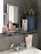 A bathroom shelf holding various skincare products, including bottles and tubes labeled with text like 'JAPANFUSION' and 'RETINOL'. A metal-framed mirror stands beside a potted green plant in a blue ceramic pot. The sink below features a chrome faucet with vintage-style handles, and a bar of soap is placed beside the skincare items.