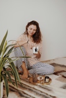 A woman wearing a striped yellow and white top with blue and white pajama pants sits comfortably on a soft, patterned blanket. She is holding a mug and smiling softly, appearing relaxed. In front of her, there is a wooden tray with a small vase, a candle, and a few decorative items. Next to her is a green houseplant.