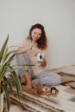 A woman wearing a striped yellow and white top with blue and white pajama pants sits comfortably on a soft, patterned blanket. She is holding a mug and smiling softly, appearing relaxed. In front of her, there is a wooden tray with a small vase, a candle, and a few decorative items. Next to her is a green houseplant.