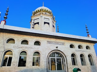 A beautifully designed mosque with a prominent dome and intricate architectural details. The structure features multiple arched windows and inscribed calligraphy above the entrance. The sky is clear and blue, enhancing the mosque's architectural elegance.