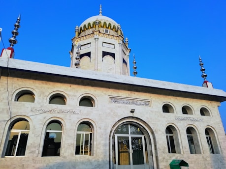 A beautifully designed mosque with a prominent dome and intricate architectural details. The structure features multiple arched windows and inscribed calligraphy above the entrance. The sky is clear and blue, enhancing the mosque's architectural elegance.