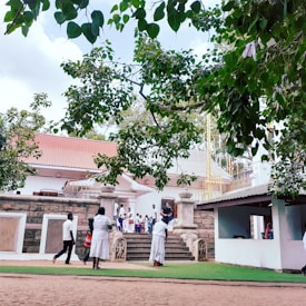 A group of people dressed in traditional clothing are gathered near an ornate building with a tiled roof and stone steps. Large green leaves from a tree hang prominently in the foreground, casting shade over the scene. The background includes architectural elements with white trim and some figures are seen interacting or walking by.