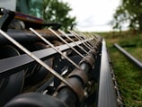 The image captures a close-up view of agricultural machinery, possibly a combine harvester or similar equipment, with sharp metal components aligned in parallel. The background features blurred greenery, hinting at a rural or farm setting.