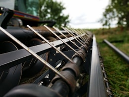 The image captures a close-up view of agricultural machinery, possibly a combine harvester or similar equipment, with sharp metal components aligned in parallel. The background features blurred greenery, hinting at a rural or farm setting.