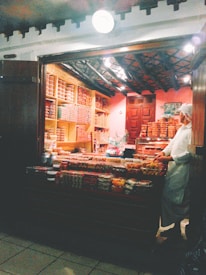 A small, cozy shop with wooden shelves and counters displaying a variety of food items. The shop is dimly lit with a warm ambiance, featuring jars, containers, and trays filled with what appear to be sweets or snacks. A person wearing a white outfit and cap is standing behind the counter, organizing or selling the items.