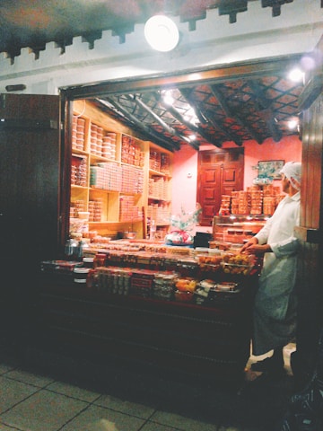 A small, cozy shop with wooden shelves and counters displaying a variety of food items. The shop is dimly lit with a warm ambiance, featuring jars, containers, and trays filled with what appear to be sweets or snacks. A person wearing a white outfit and cap is standing behind the counter, organizing or selling the items.