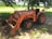 A bright orange Kubota tractor is parked on a patch of dirt under the shade of a tree. The tractor has large, rugged tires and a front loader attachment with a visible scoop. In the background, there is a field of corn and a small road. Sunlight filters through the leaves of the tree, casting shadows on the tractor and the ground.