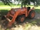 A bright orange Kubota tractor is parked on a patch of dirt under the shade of a tree. The tractor has large, rugged tires and a front loader attachment with a visible scoop. In the background, there is a field of corn and a small road. Sunlight filters through the leaves of the tree, casting shadows on the tractor and the ground.