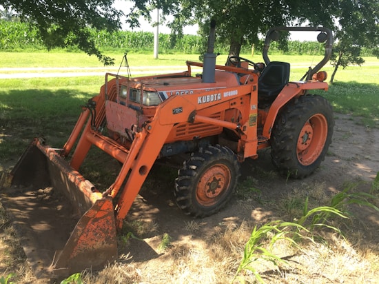 A bright orange Kubota tractor is parked on a patch of dirt under the shade of a tree. The tractor has large, rugged tires and a front loader attachment with a visible scoop. In the background, there is a field of corn and a small road. Sunlight filters through the leaves of the tree, casting shadows on the tractor and the ground.