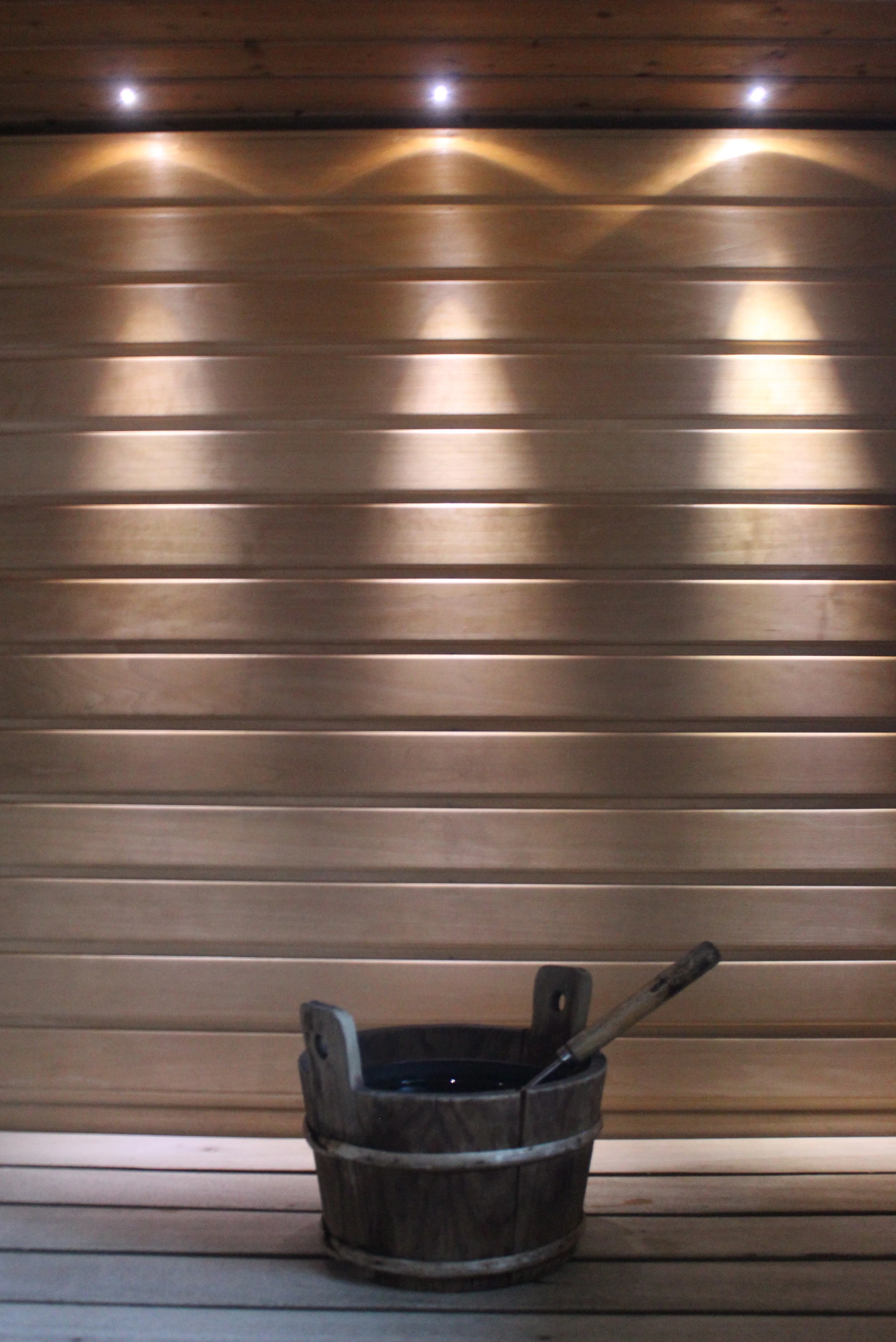 Horizontal timber slats lit by warm downlights above a wooden sauna bucket and ladle