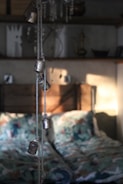 A softly lit bedroom interior with rustic elements. In the foreground, several small metal bells hang from a rope, casting gentle shadows on the wall behind. The bed features patterned bedding with a mix of colors, adding a cozy and inviting feel. Shelves in the background hold various decorative items, including a candlestick and some abstract art pieces, contributing to the quaint and eclectic atmosphere.