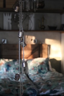 A softly lit bedroom interior with rustic elements. In the foreground, several small metal bells hang from a rope, casting gentle shadows on the wall behind. The bed features patterned bedding with a mix of colors, adding a cozy and inviting feel. Shelves in the background hold various decorative items, including a candlestick and some abstract art pieces, contributing to the quaint and eclectic atmosphere.