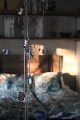 A softly lit bedroom interior with rustic elements. In the foreground, several small metal bells hang from a rope, casting gentle shadows on the wall behind. The bed features patterned bedding with a mix of colors, adding a cozy and inviting feel. Shelves in the background hold various decorative items, including a candlestick and some abstract art pieces, contributing to the quaint and eclectic atmosphere.