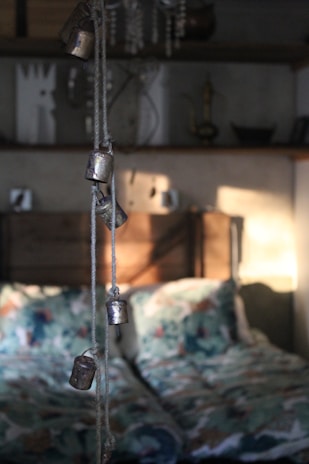 A softly lit bedroom interior with rustic elements. In the foreground, several small metal bells hang from a rope, casting gentle shadows on the wall behind. The bed features patterned bedding with a mix of colors, adding a cozy and inviting feel. Shelves in the background hold various decorative items, including a candlestick and some abstract art pieces, contributing to the quaint and eclectic atmosphere.