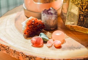 Several polished and raw crystals are arranged on a wooden surface. The crystals vary in color, including red, purple, green, and pink, and have different shapes and sizes. The background features a glass container with golden speckles and a soft, warm lighting that highlights the textures of the wood and stones.