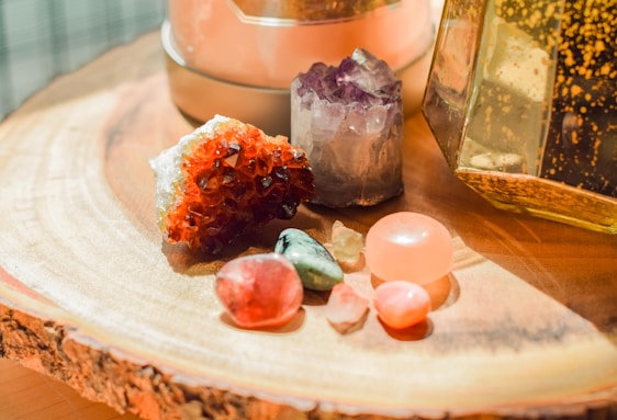 Several polished and raw crystals are arranged on a wooden surface. The crystals vary in color, including red, purple, green, and pink, and have different shapes and sizes. The background features a glass container with golden speckles and a soft, warm lighting that highlights the textures of the wood and stones.