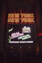 A neon sign displaying 'New York New York' with bright red letters. Below it, a digital screen advertises a real coaster with virtual thrills and suggests purchasing tickets inside. People are visible in shadow in front of the sign.
