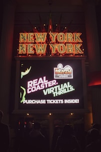 A neon sign displaying 'New York New York' with bright red letters. Below it, a digital screen advertises a real coaster with virtual thrills and suggests purchasing tickets inside. People are visible in shadow in front of the sign.