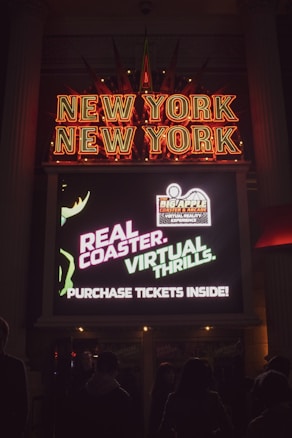 A neon sign displaying 'New York New York' with bright red letters. Below it, a digital screen advertises a real coaster with virtual thrills and suggests purchasing tickets inside. People are visible in shadow in front of the sign.