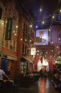 A bustling indoor street setting with brick buildings and various shop signs, illuminated by string lights overhead. People are seated at outdoor tables, enjoying food and conversation, while others stand or walk by. The ambiance is warm and inviting, with a mix of vintage and urban elements.
