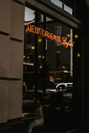 A dimly lit street scene is reflected in a large window, featuring a neon sign that reads 'New Orleans Seafood.' A person is sitting inside, partially visible through the window among reflections of other buildings and street activities.