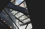 A modern architectural interior with geometric patterns created by large glass panels and metal beams. The view includes an exterior urban skyline with various high-rise buildings visible through the glass. The composition plays with light and shadow, enhancing the contrasting shapes and textures.