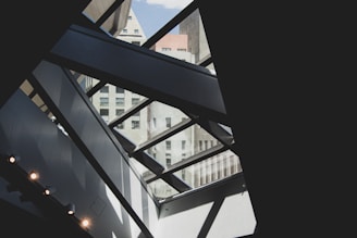 A modern architectural interior with geometric patterns created by large glass panels and metal beams. The view includes an exterior urban skyline with various high-rise buildings visible through the glass. The composition plays with light and shadow, enhancing the contrasting shapes and textures.