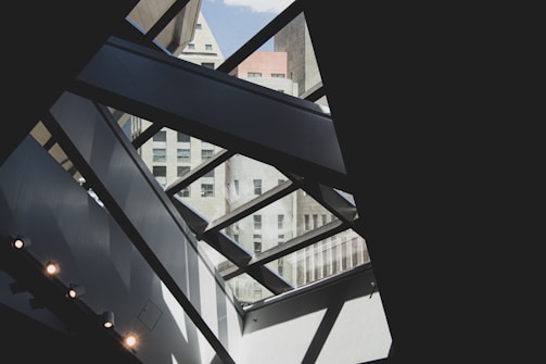 A modern architectural interior with geometric patterns created by large glass panels and metal beams. The view includes an exterior urban skyline with various high-rise buildings visible through the glass. The composition plays with light and shadow, enhancing the contrasting shapes and textures.