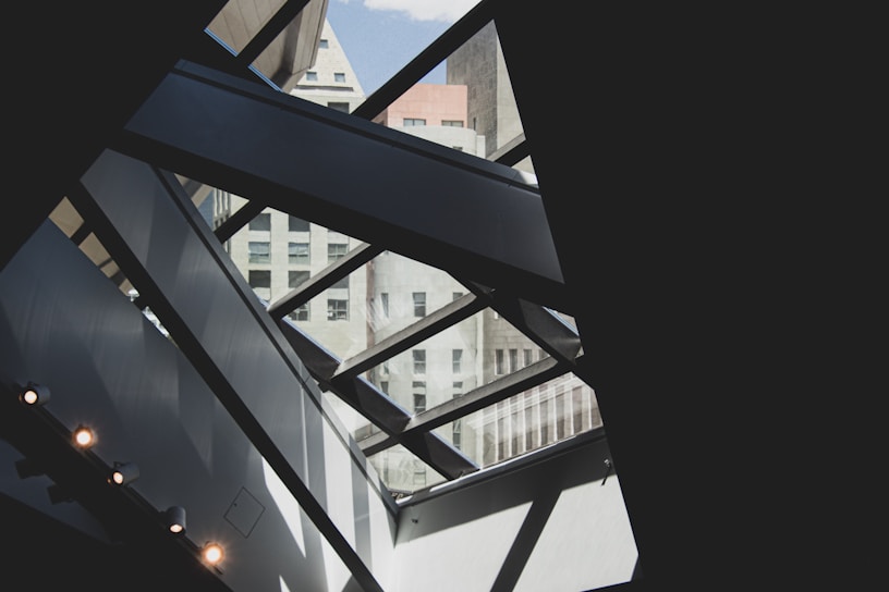 A modern architectural interior with geometric patterns created by large glass panels and metal beams. The view includes an exterior urban skyline with various high-rise buildings visible through the glass. The composition plays with light and shadow, enhancing the contrasting shapes and textures.