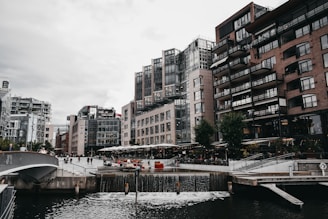 Urban waterfront scene featuring modern buildings with extensive glass facades and brick construction. A small waterfall flows into a body of water in the foreground, surrounded by a walkway and bridge. People are leisurely walking or sitting by outdoor cafes lined with greenery.
