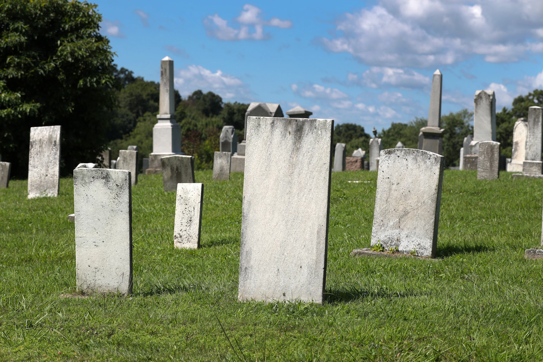 Plain white marble slabs and obelisks on a mown hillside under a bright blue midday sky, their faces washed flat by overhead light