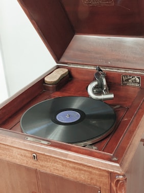An antique wooden phonograph with a black vinyl record on its turntable. The phonograph features a classic speaker horn and has a label that reads 'Victor' on the interior. The device is open, displaying its intricate mechanical parts and a cloth on the side.