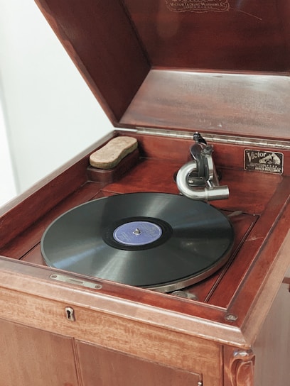 An antique wooden phonograph with a black vinyl record on its turntable. The phonograph features a classic speaker horn and has a label that reads 'Victor' on the interior. The device is open, displaying its intricate mechanical parts and a cloth on the side.