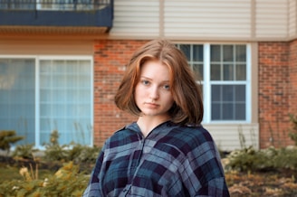 A person with short, brown hair stands outside in front of a house with brick and siding. The person is wearing a plaid shirt and has a neutral expression. The background includes some shrubbery and a window.
