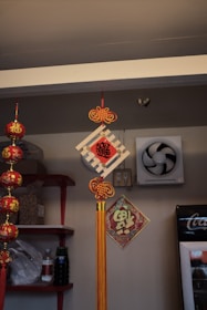 Hanging decorations featuring traditional Chinese motifs are displayed indoors. Red and gold adornments with Chinese characters are suspended, accompanied by a red and gold tassel. Shelves with bottles and bags are visible in the background, alongside a wall fan and a refrigerator with visible branding.